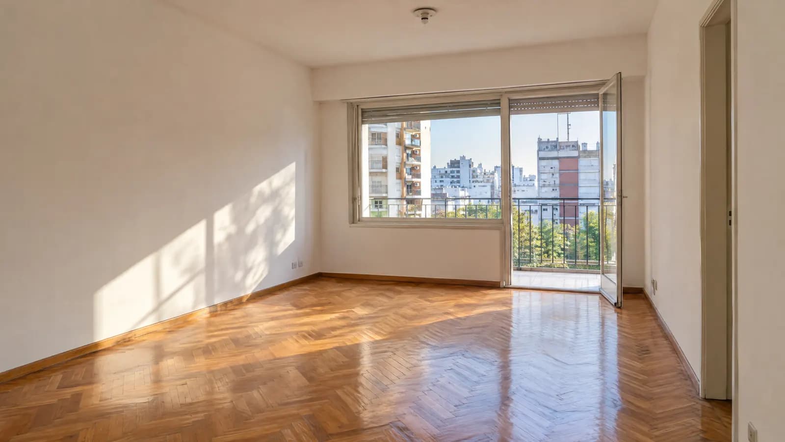 Living vacío y luminoso de un departamento con piso de parquet y ventanal al balcón.
