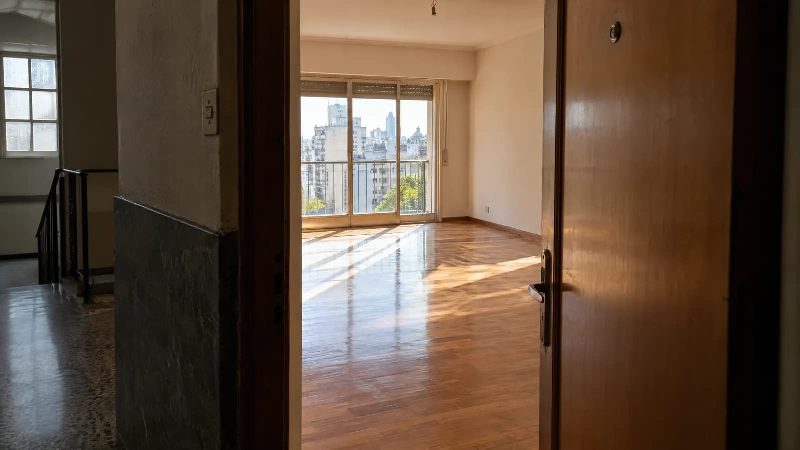 Puerta abierta de un departamento que deja ver un living vacío con luz natural.