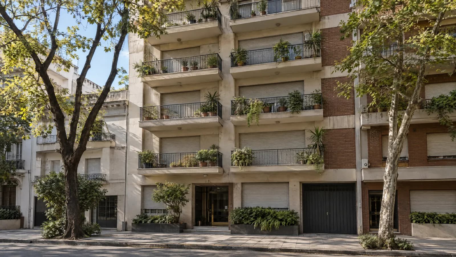 Fachada de un edificio de departamentos con balcones y plantas en una calle arbolada de Buenos Aires.
