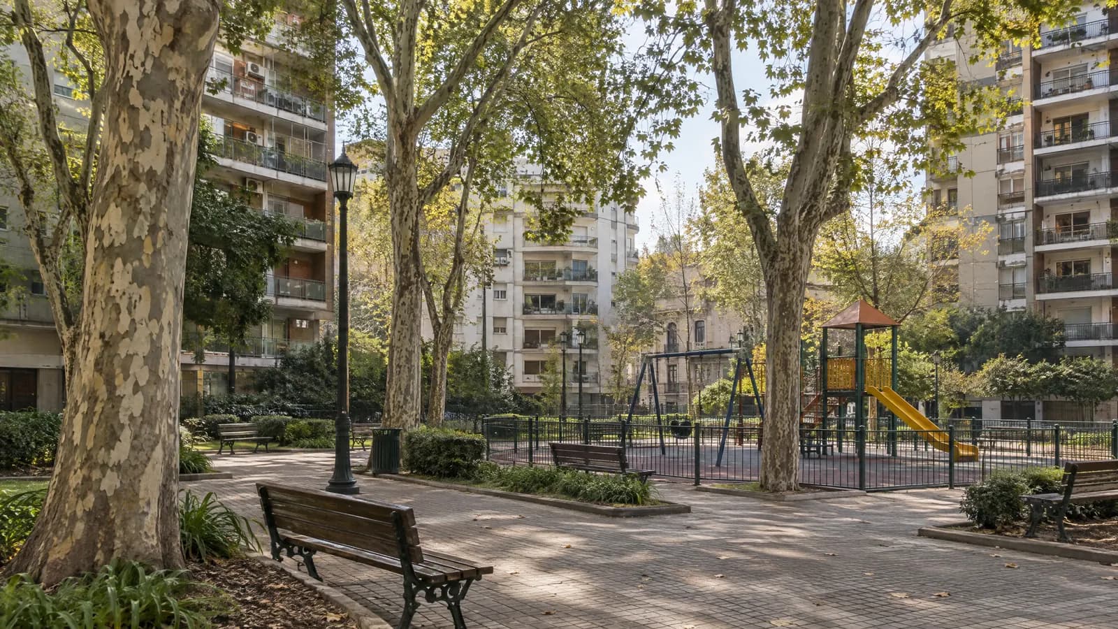 Plaza de barrio con árboles, bancos y juegos, rodeada de edificios.