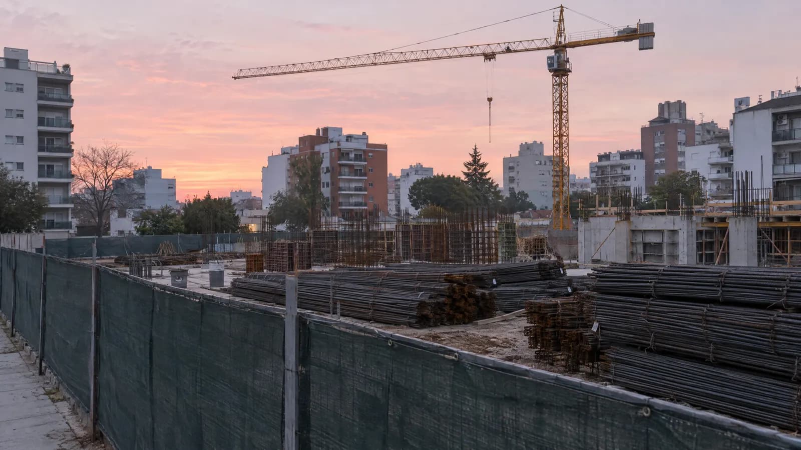 Obra cercada con acopio de hierros y una grúa al amanecer.
